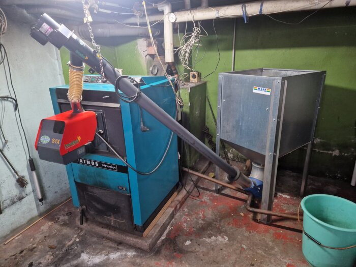 Pelletsbrännare av märket Atmos i ett rum med grön vägg och kringliggande utrustning, inklusive en metalltunna och en turkos plasthink.