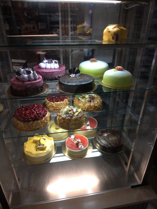 Olika tårtor och bakverk i en glasmonter på ett café, bland annat chokladtårta, prickig rosa tårta och grön prinsesstårta med blommor.