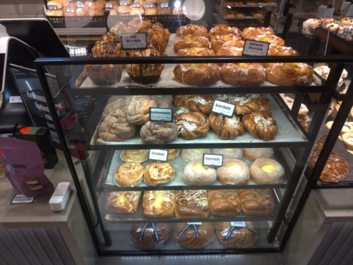 Bageridisk med olika bakverk som kanelbullar, wienerbröd och andra sötsaker uppställda i rader med prislappar i ett kafé.