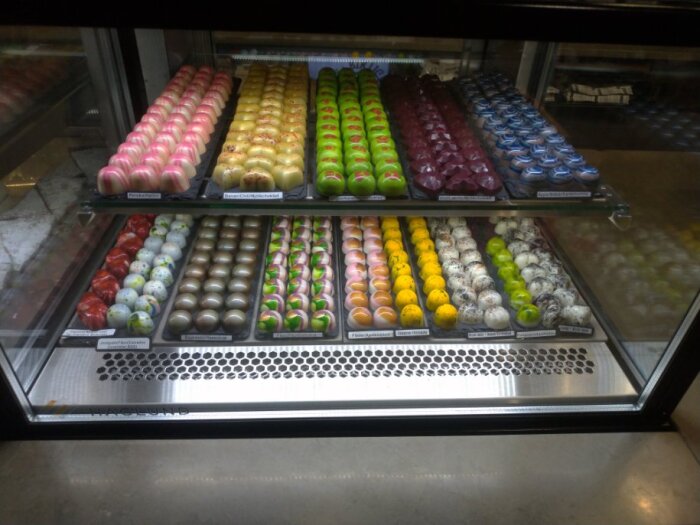 Praliner i olika färger och smaker uppställda i ett glasmonter.