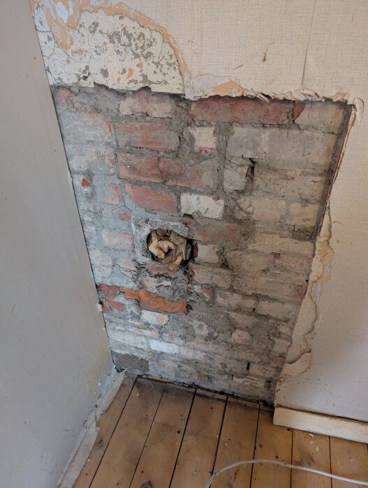 Gammalt tegel med hål fylld med tidningspapper, del av murstock från 1907 i behov av renovering. Sliten vägg och golv synliga.