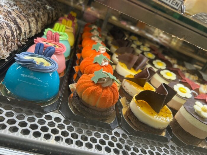 Ett urval av färgglada desserter och bakverk i en glasmonter på ett konditori, med olika dekorationer och smaker.
