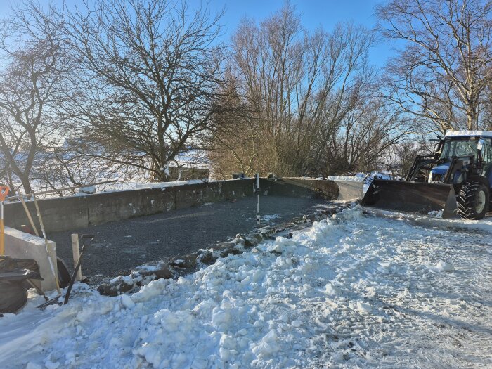 Snötäckt byggplats med traktor bredvid betonggrund för ett växthus i södra Skåne. Kullerstenar syns längs kanten. Träd i bakgrunden.