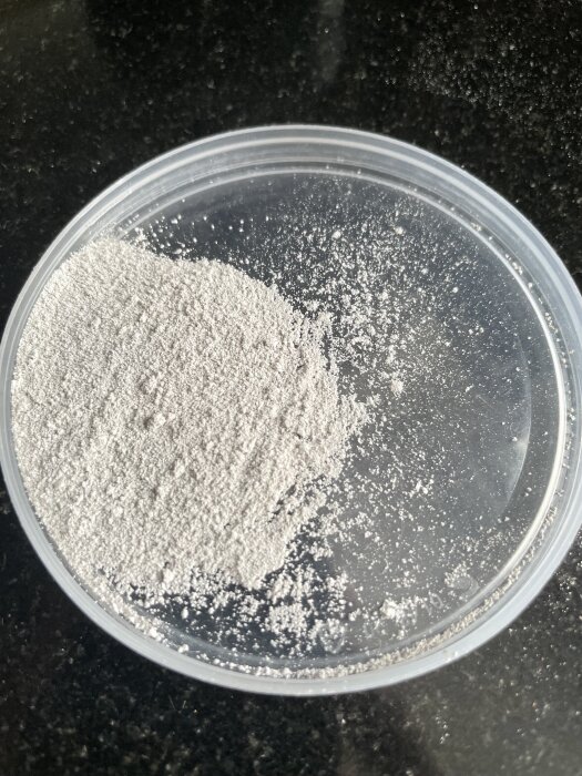 Grå sand- eller stenliknande pulver i en plastbehållare på en mörk yta.