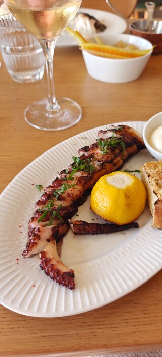 Grillad bläckfiskarm med citron, garnerad med grönt, serverad på en tallrik med bröd vid sidan, och ett glas vin i bakgrunden.