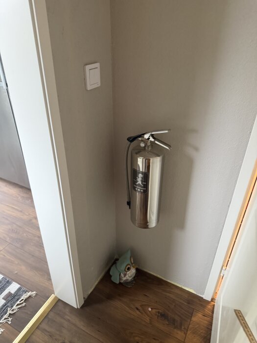 Brandsläckare monterad på väggen i ett hörn, med en liten ugglefigur på golvet intill.