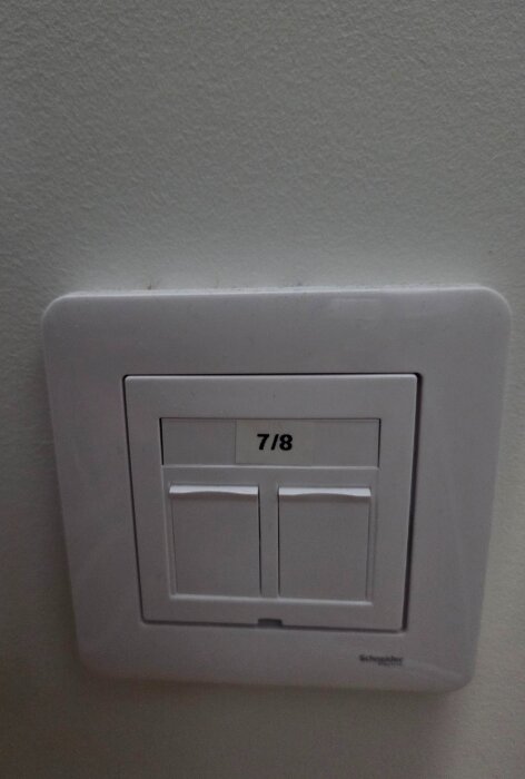 Dubbla vita ljusströmbrytare monterade på en vägg, märkta med "7/8", tillverkarens logotyp synlig längst ner.