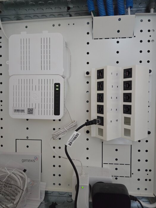 Internet- och nätverksutrustning installerad på en vit perforerad vägg med kablar och portar, inklusive en Genexis-enhet och nätverksanslutningar.