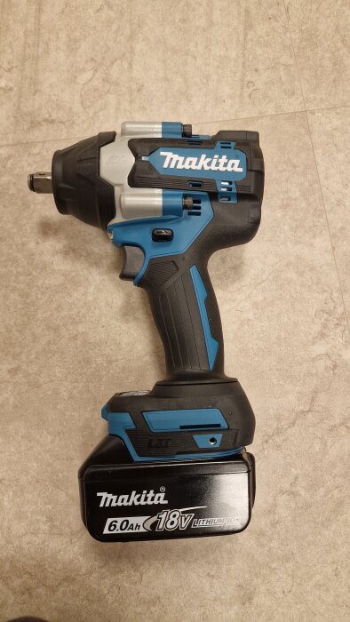 Blå och svart Makita batteriborrmaskin, 18V, med 6.0Ah Litiumjonbatteri, liggande på en grå klinkergolv.