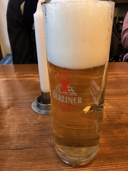 Ett glas öl med mycket skum, märkt "Berliner", står på ett träbord bredvid ett tänd ljus.