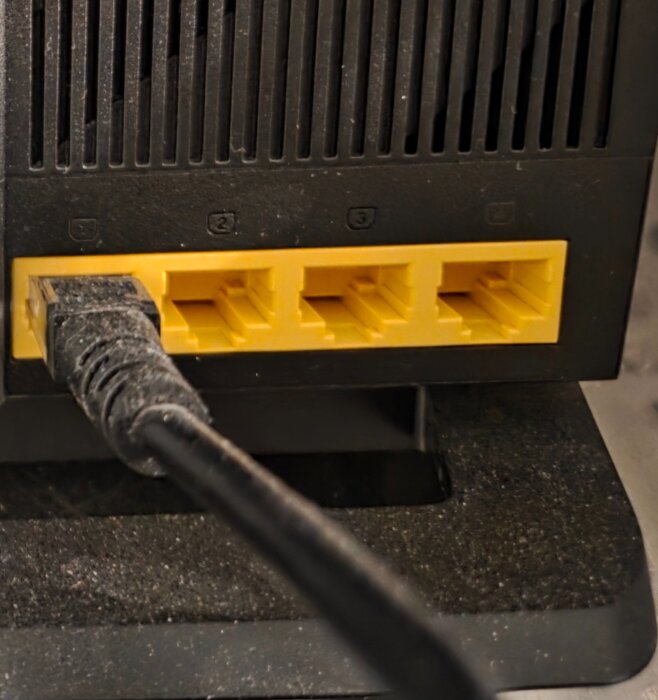 Svart nÀtverkskabel ansluten till en av de fyra gula Ethernet-portarna pÄ en router.