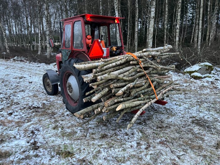 Röd traktor i en skogsområde, lastad med ved på marken täckt av lätt snö.