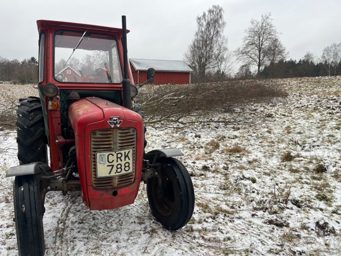 Röd traktor med registreringsskylt på en snötäckt åker, omgiven av grenar. I bakgrunden syns ett rött hus och en lada.