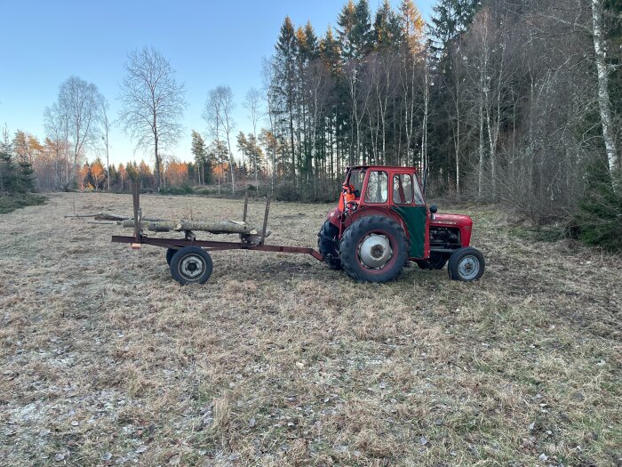 Röd traktor med släpkärra fylld med ved på ett öppet fält, omgivet av skog.