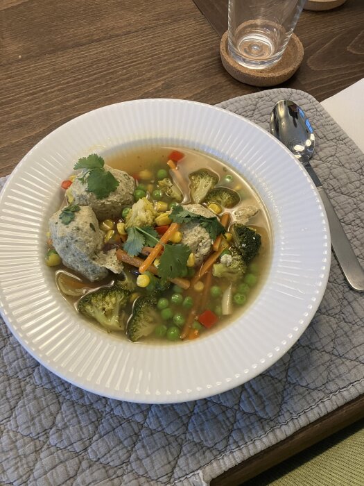 Kycklingfrikadeller i buljong med broccoli, ärter, morot och koriander i en vit tallrik på ett grått bordstabletter.