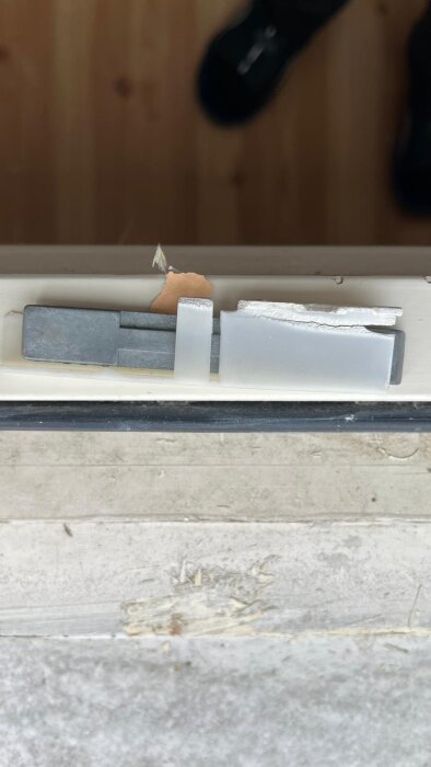 Närbild av trasigt fönsterbeslag i plast och metall, med skadad yta runt om.