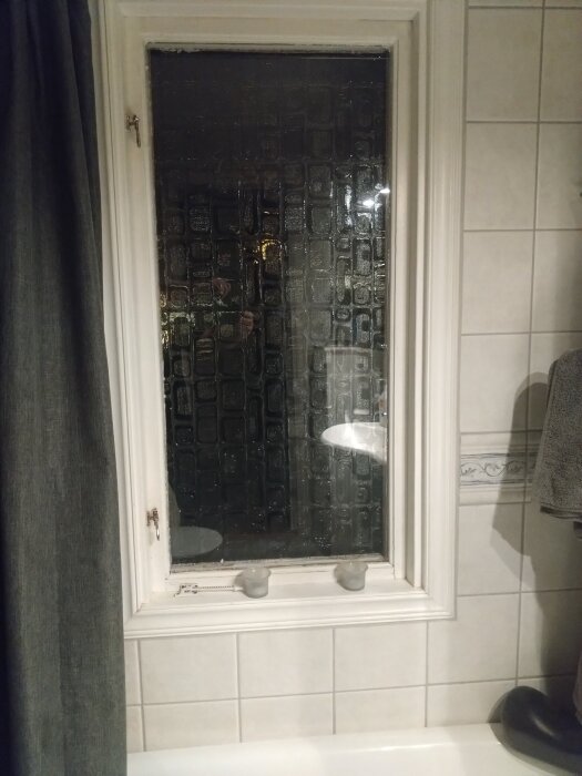 Fönster i badrum med fodrar och kakel runtom, insatt ljuslykta på fönsterbrädan.