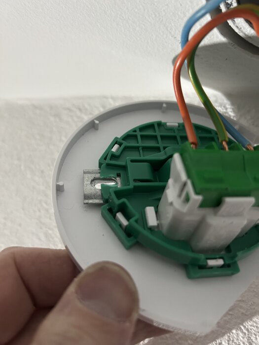 Hand håller i ett elektriskt uttag; grönt och vitt inre med kablar, och synliga metallklor som blockerar installation.