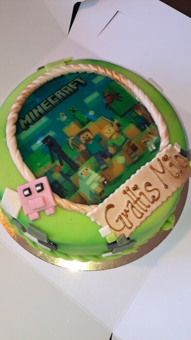 Grön tårta med ett Minecraft-motiv och figurer, samt texten "Grattis Mio" på ett dekorativt band.