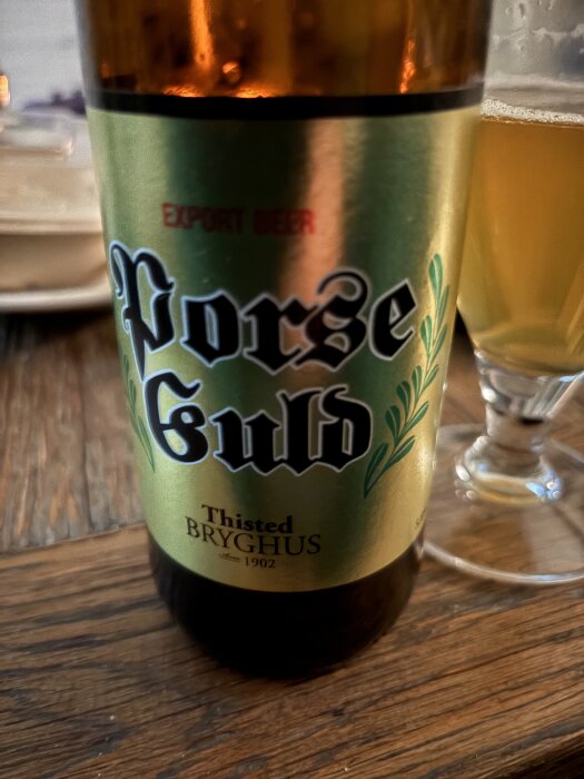 Flaska av Thisted Bryghus Porse Guld lager på ett träbord, delvis med ett glas öl bredvid.