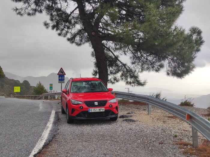 Röd bil parkerad vid en trafikerad bergsväg med varningsskylt för kurvor, ett träd och en vy över berg i bakgrunden.