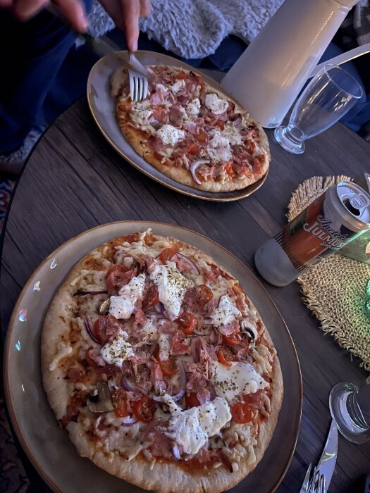 Två pizzor på tallrikar med skinka, tomater och ost, en citrusläsk och en vit kanna på ett mörkt träbord.