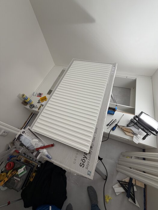 Målat vitt element på arbetsyta med målarverktyg och material i ett stökigt rum under renovering.