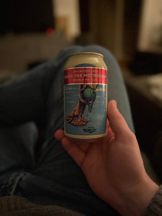 En hand håller en ölburk med etiketten "Radio the Mothership" från Collective Arts, Double IPA. En person sitter avslappnat i bakgrunden.