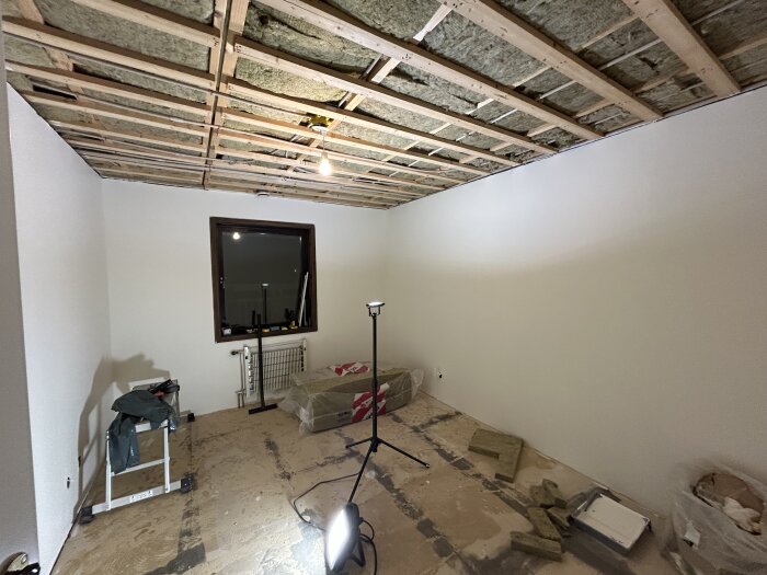 Under renovering av rum med synligt träreglat tak, isolering, stege och byggmaterial på golvet. Fönster och belysning i rummet.
