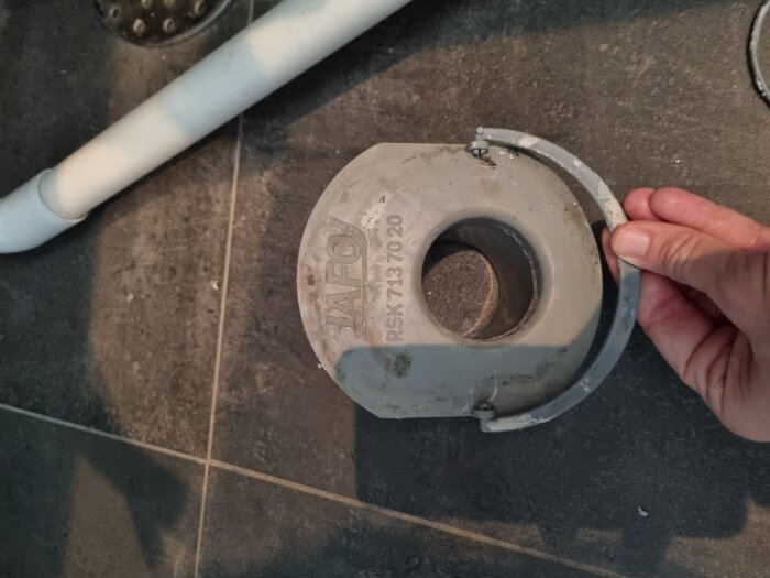 En hand håller en grå golvbrunnsinsats med märket "JAFO" och ett avtagbart tätningsring, på ett mörkt kakelgolv bredvid ett vitt rör.