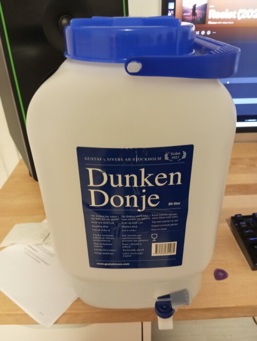 En 20-liters plastdunk med blå lock och handtag, märkt "Dunken Donje", står på ett bord nära en dator.