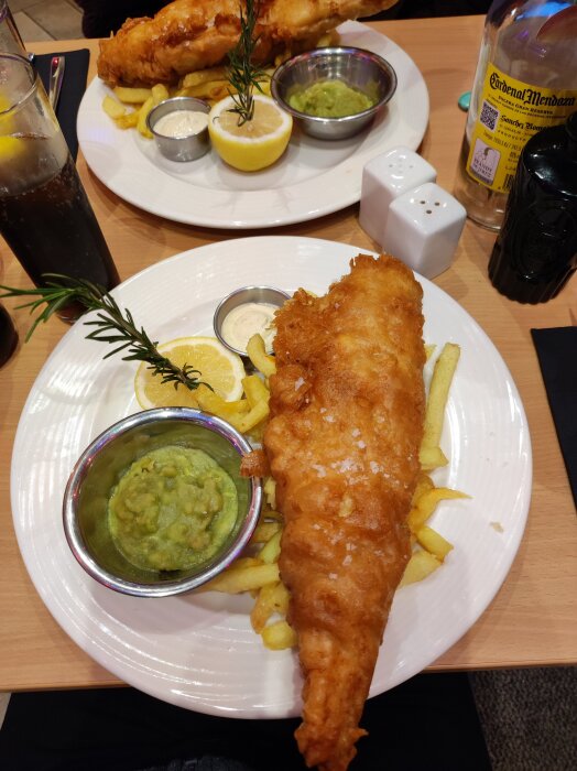 Fish and chips serverade med mosad ärta, citronhalvor, sås och en kvist rosmarin på en vit tallrik, tillsammans med en flaska dryck.