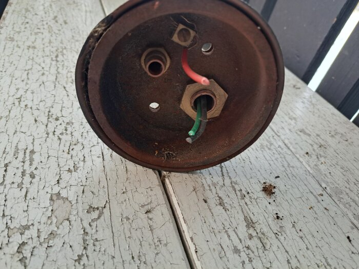 Rostig metallbehållare med tre elektriska kablar, stående på ett ytskikt med flagnande vit färg.