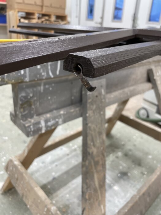 Brunt trä med ett metallbeslag på änden, troligen del av fönsterspröjs, med suddig bakgrund av träverkstad.