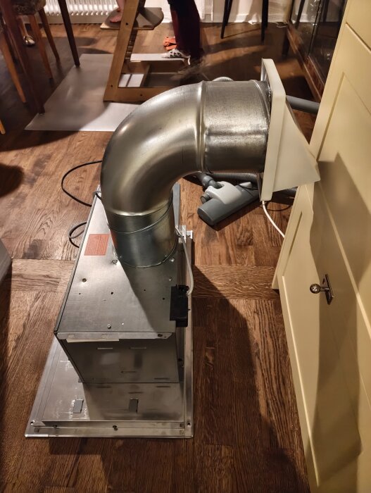 Ventilationssystem med spirorör och brandspjäll installerat ovanför kylskåp på trägolv i kök, med synlig 90-graders böj.
