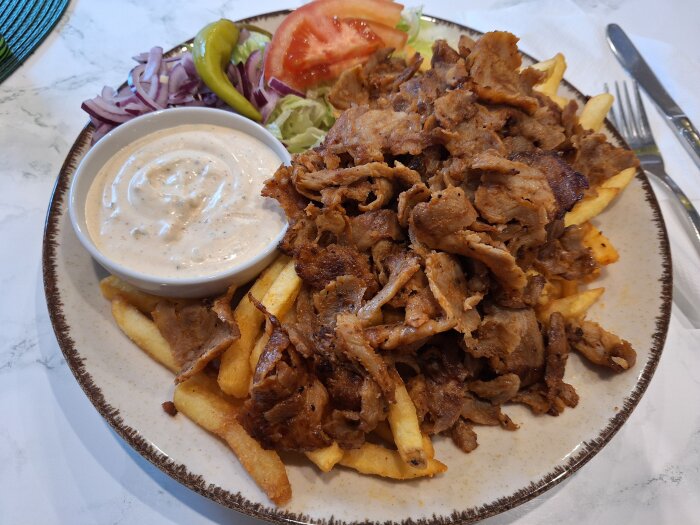 Tallrik med kebabkött och pommes frites, serverad med vitlöksdressing, skivad tomat, paprika och rödlök som tillbehör.
