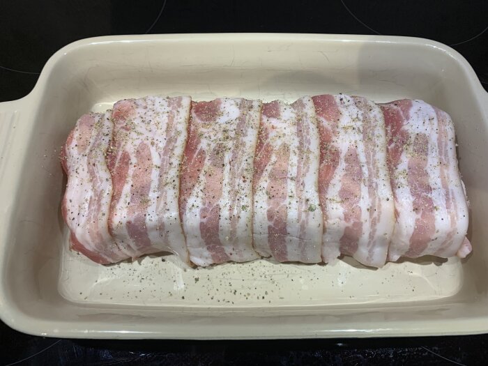 Köttfärslimpa lindad i bacon, kryddad och redo för ugnsbakning i en vit form.