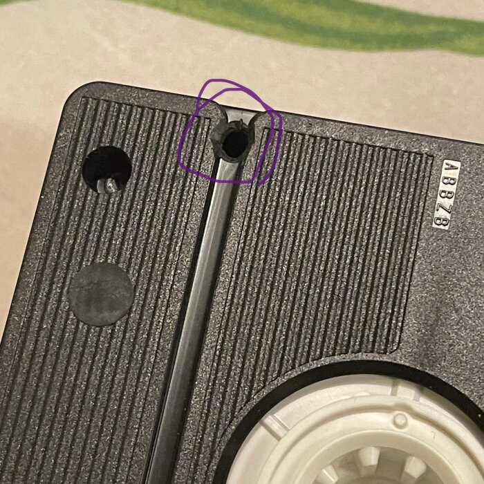 Närbild av ett svart VHS-band med en plastbit markerad med en lila cirkel. Bakgrunden är suddig.
