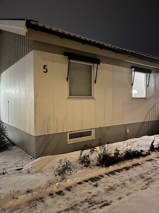 En enplansvilla från 70-talet med snö på marken, liggande fjällpanel och eternitskivor längst ner, husnummer 5 är synligt.