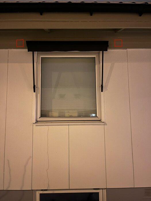 Fasad med liggande fjällpanel ovanför fönster, två röda markeringar nära taket.