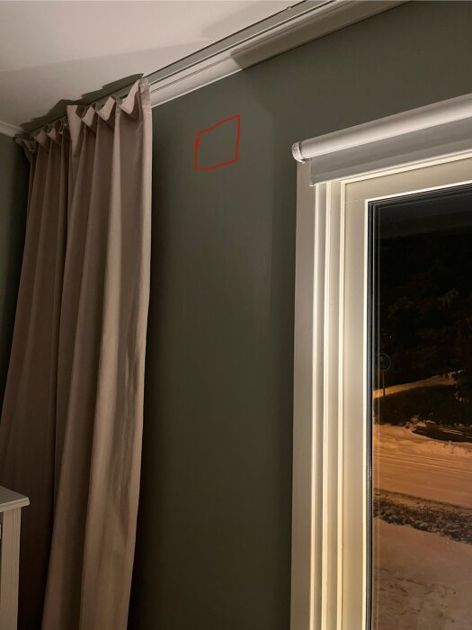 Vardagsrumsvägg med rödmarkerad ruta nära taket, bredvid ett fönster med persienn och ljusa gardiner.