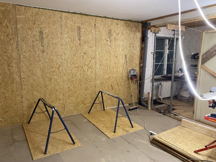 Rum under renovering med OSB-skivor på golvet och väggen. Lutning synlig med temporär uppställning och sladdar. Bygglampor och verktyg i bakgrunden.