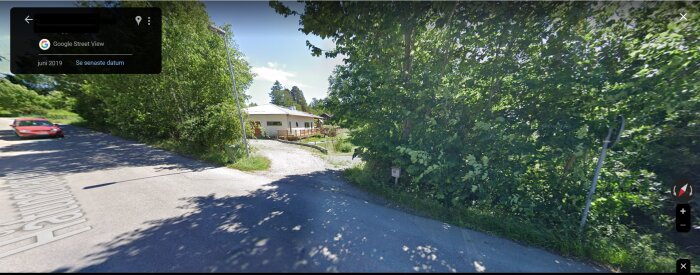 En uppfart med grus som leder till ett hus omgivet av gröna träd och buskar, med en asfalterad väg i framkant.