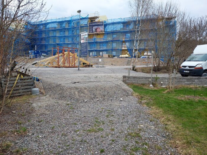 Infart med grus och gräsmatta i förgrunden, byggarbetsplats i bakgrunden med blått skynke och en vit skåpbil till höger.
