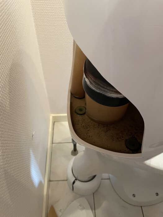 Baksidan av en toalett med avtaget bakstycke, visar rör och beslag. Toaletten står på ett kaklat golv intill en ljus vägg.