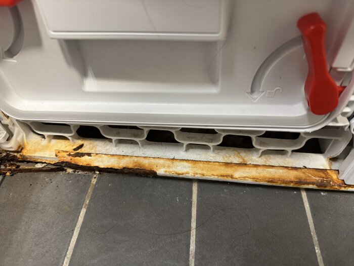 Rostfläckar på botten av en vit torktumlare, nära golvet, med en detalj av en röd knapp eller spak.