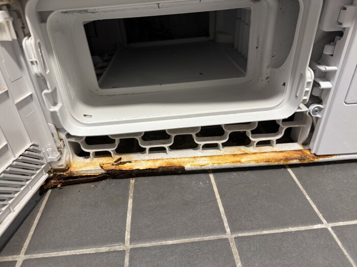 Rostskador på botten av en Cylinda torktumlare, nära vänstra hörnet, inomhus i ett badrum med grått kakelgolv.