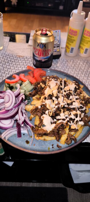 Tallrik med kebabkött, pommes frites och sås, omgiven av gurka, tomat och rödlök. En burk Norrlands Guld öl och kryddflaskor i bakgrunden.