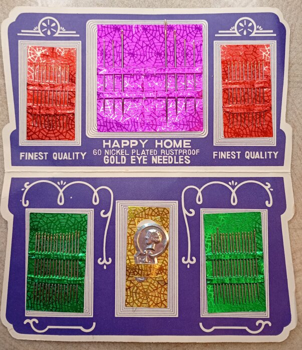 Förpackning med "Happy Home" synålar i olika färger: röd, lila och grön. Texten beskriver dem som "finest quality" och "rustproof gold eye needles".