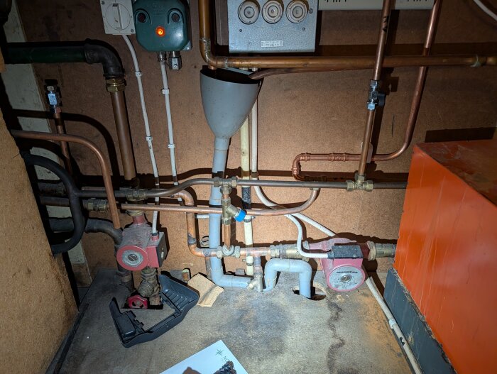 Värmesystem i äldre hus med rör, ventiler och en orange elpanna. Elkabel och rördragning syns i ett trångt utrymme.